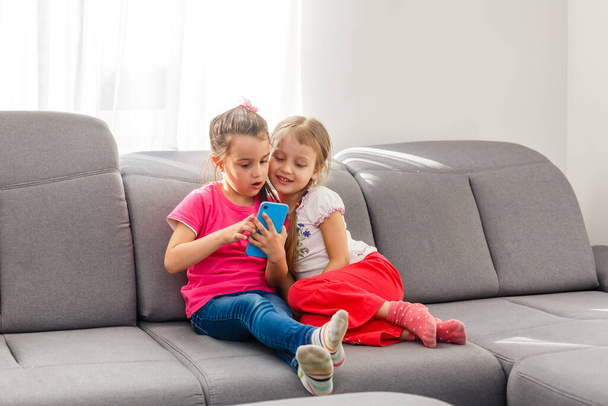 Stay Home Stay Safe. Two happy kids sitting on cozy chair relaxing playing in white living room indoors. Sisters having fun at home - Фото, зображення
