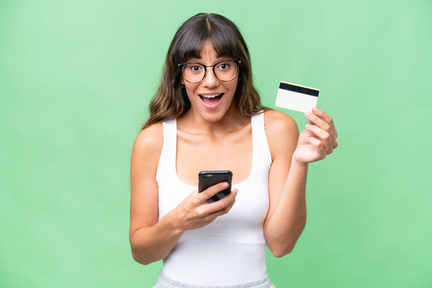Young caucasian woman over isolated background buying with the mobile and holding a credit card with surprised expression - Foto, imagen