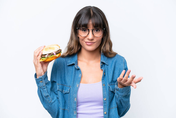 Joven mujer caucásica sosteniendo una hamburguesa aislada sobre fondo blanco haciendo gestos de dudas mientras levanta los hombros - Foto, imagen