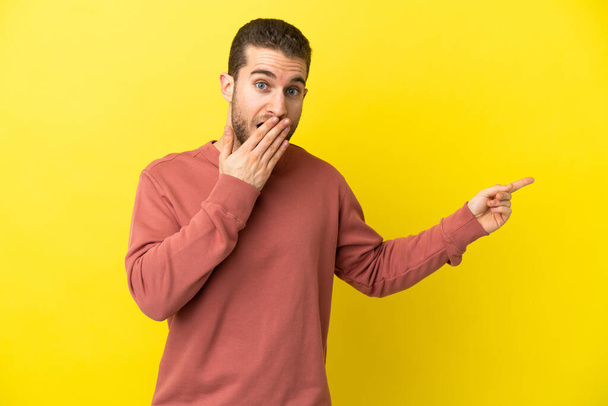 Handsome blonde man over isolated yellow background with surprise expression while pointing side - Foto, immagini