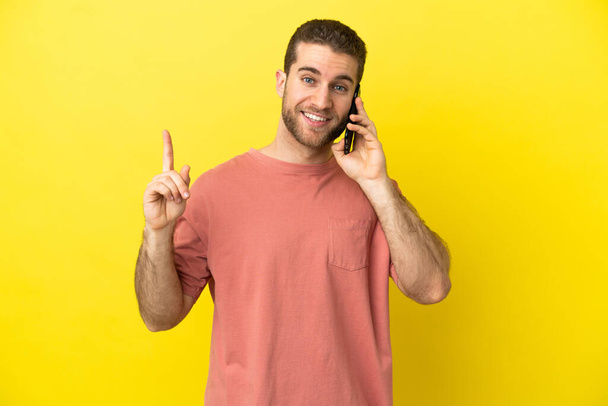 Yakışıklı sarışın adam izole edilmiş arka planda cep telefonu kullanıyor ve en iyinin işareti olarak parmağını kaldırıyor. - Fotoğraf, Görsel