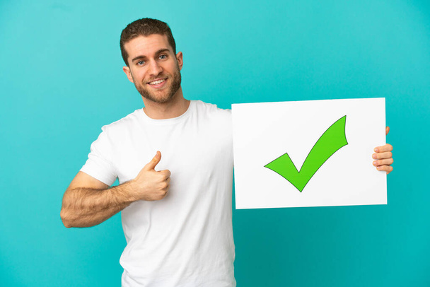 Hezké blondýny muž přes izolované modré pozadí drží plakát s textem Green check mark ikona s palcem nahoru - Fotografie, Obrázek