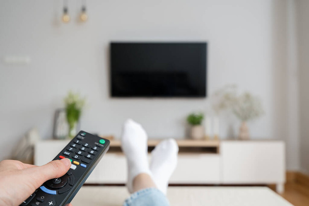 ženská ruka držet TV dálkové ovládání a stiskněte tlačítko pro zapnutí televize, posezení na gauči v obývacím pokoji v útulném bytě, zábavu a relaxaci doma - Fotografie, Obrázek