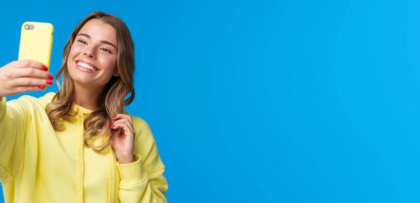 Close-up portrait tender cute blond girl in yellow hoodie, holding mobile phone, taking selfie with smartphone posing adding photo filter in appllication, standing blue background. - Photo, Image
