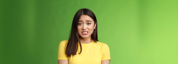 Anxious worried cute asian girl brunette clench teeth frowning shaking from fear look guilty afraid, stand nervous awaiting punishment, green background, apologizing ashamed. Copy space - Foto, Imagen