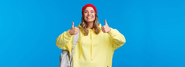 Klinkt goed, deal. Vrolijk gelukkig en glimlachend hippe meisje in rode muts en gele hoodie, goedkeuren idee, akkoord met vrienden kiezen waar hangen na de les, houden rugzak, blauwe achtergrond. - Foto, afbeelding
