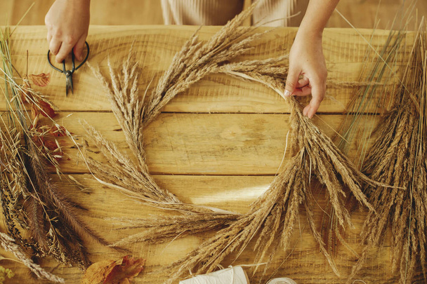 Making stylish autumn wreath on rustic table. Hands arranging dried grass on hoop on wooden table with scissors, thread,  top view. Fall decor and arrangement in farmhouse. - Fotoğraf, Görsel