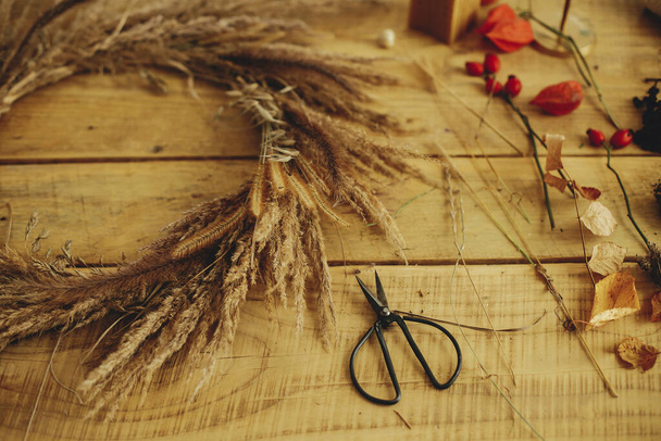 Stylish autumn wreath with dried grass, scissors, berries on wooden table. Making stylish autumn wreath on rustic table. Fall decor and arrangement in farmhouse. - Photo, Image