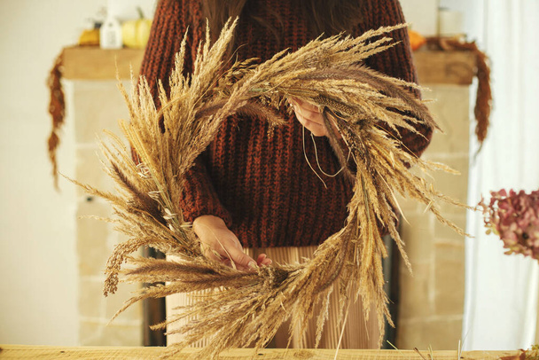 Kobieta trzymająca jesienny wieniec w rustykalnym pokoju przy kominku. Stylowy jesienny wieniec z suszoną trawą i ziołami w rękach kobiet. Jesienny wystrój i aranżacja w wiejskim domu - Zdjęcie, obraz