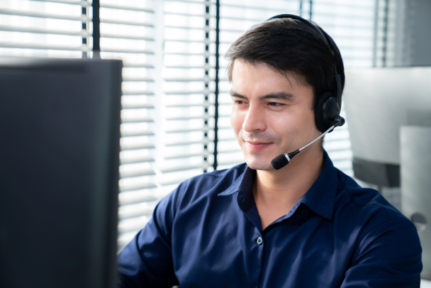 Young competent asian male call center agent working at his computer while simultaneously speaking with customers. Concept of an operator, customer service agent working in the office with headset. - 写真・画像