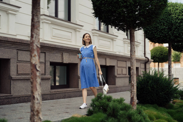 Hermosa joven embarazada en un vestido azul caminando por las calles de la ciudad tomando una copa para llevar. - Foto, Imagen