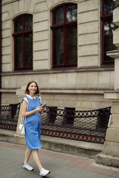 Belle jeune femme enceinte en robe bleue se promenant dans les rues de la ville à l'aide d'un smartphone. - Photo, image
