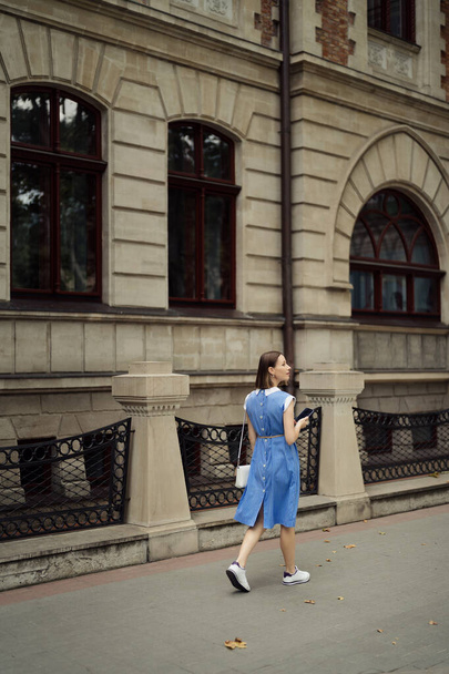 Beautiful young pregnant woman in a blue dress walking through the city streets using a smartphone. - Photo, Image