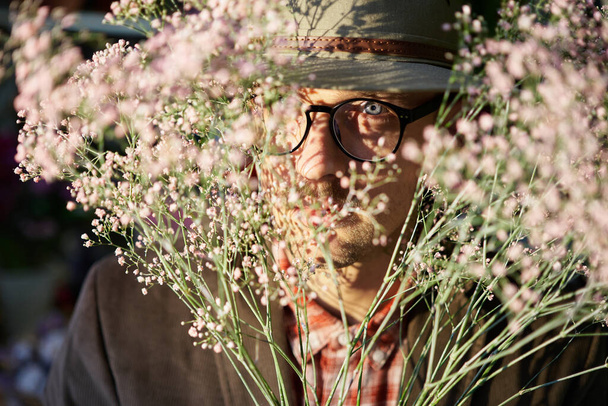 Fresh spring flowers. Cheerful male person wearing eyeglasses and hat hiding in fresh flower bouquet. Mothers Day, Valentines Day or International Womens Day concept. High quality photo - Foto, imagen