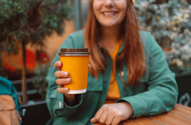 Πορτρέτο της χαμογελαστής Ευρωπαίας με κομψά φωτεινά ρούχα κάθεται με takeaway καφέ στα χέρια στο δρόμο cafe - Φωτογραφία, εικόνα