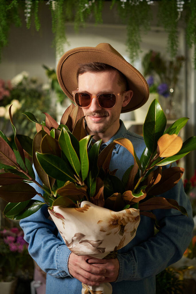Χαριτωμένο καυκάσιος hipster πρόσωπο σε γυαλιά ηλίου και καπέλο κρατώντας μεγάλο φρέσκο λουλούδι άνοιξη μπουκέτο στο ανθοπωλείο. Ημέρα της Μητέρας, Ημέρα του Αγίου Βαλεντίνου ή Διεθνής Ημέρα της Γυναίκας έννοια. Υψηλής ποιότητας εικόνα - Φωτογραφία, εικόνα