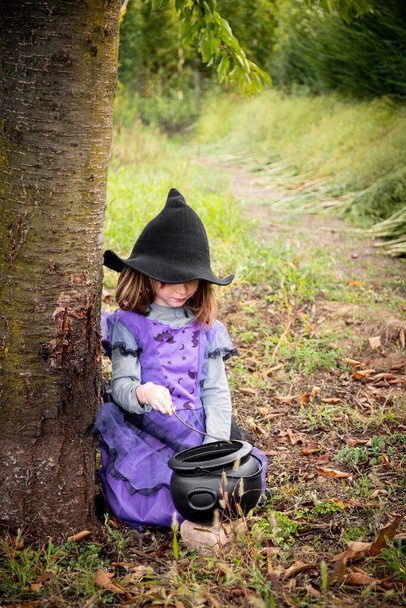 Caucasian child girl, 6 years old, in halloween purple witch costume and black hat, playing outdoor with a cauldron. Vertical shot. - Photo, Image