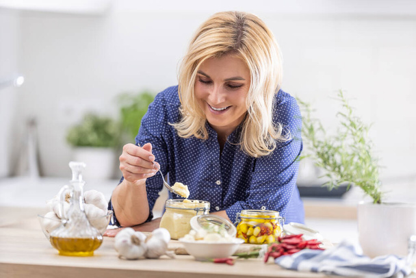 Smiling blonde woman takes spoonful of crushed garlic during preserves making session in the autumn. - 写真・画像