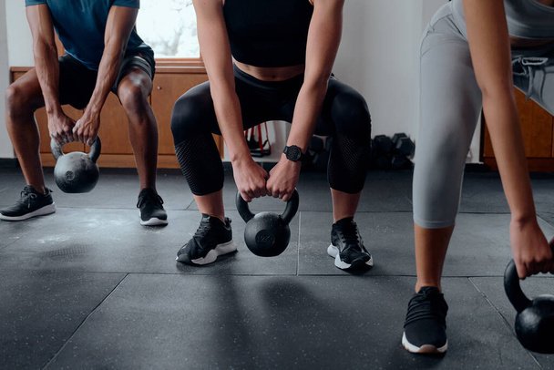 Close up of three determined multiracial young adults in sportswear doing kettlebell squats at the gym - Fotoğraf, Görsel