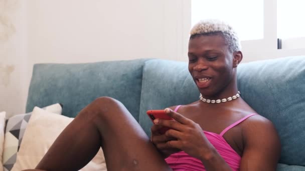 Вражений афроамериканець у стильному жіночому одязі, який пише смартфон і слухає голосове повідомлення, сидячи вдома на дивані. - Кадри, відео