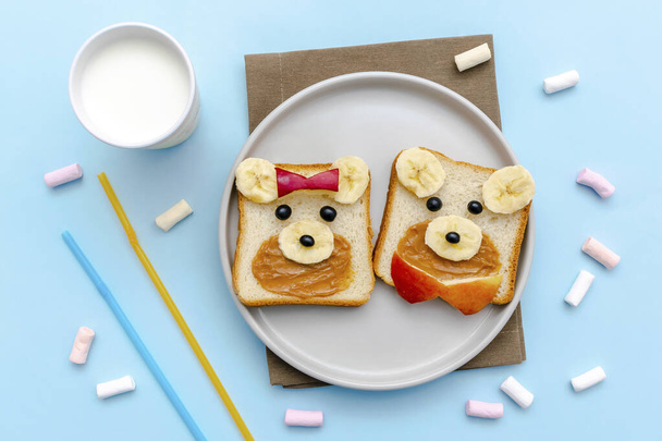 Vicces aranyos medve, kutyaarcú szendvics kenyér mogyoróvajjal, banánnal, almával, tejjel, mályvacukorral. Gyerekek gyerekek babák édes desszert egészséges reggeli ebéd élelmiszer művészet tányéron, lapos fektetés, felülnézet. - Fotó, kép