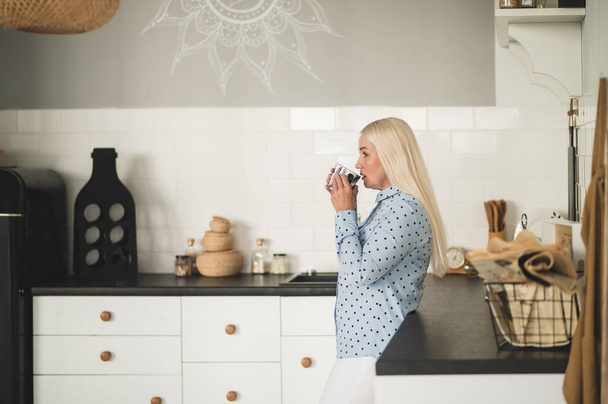Side view of a serious pensive blonde female leaning against the kitchen counter and drinking coffee - Фото, изображение