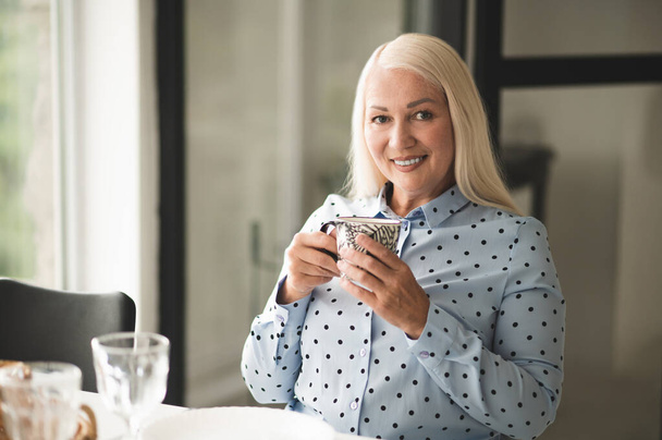 Waist-up portrait of a joyous woman with a ceramic mug in her hands sitting at the window table - Photo, image