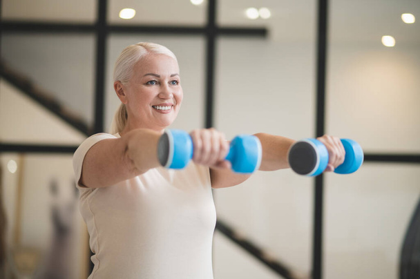 Waist-up portrait of a smiling cheerful beautiful mature blonde female doing a dumbbell front raise - Valokuva, kuva