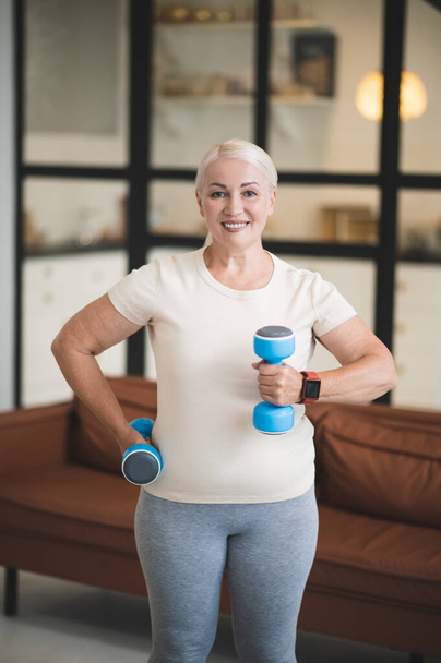 Smiling happy blonde mature Caucasian woman exercising with a pair of dumbbells in the room - Valokuva, kuva