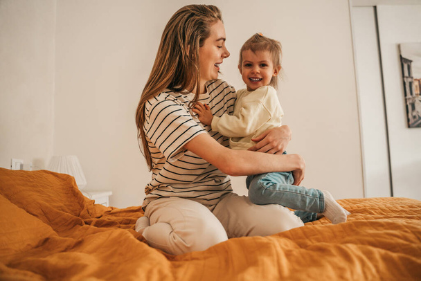 Smiling happy loving young female parent sitting on the bed and hugging her cute daughter - Fotografie, Obrázek