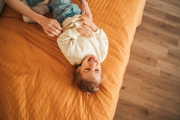 Top view of a pleased child lying on the quilted bedspread in the bedroom being tickled by her mother - Fotografie, Obrázek