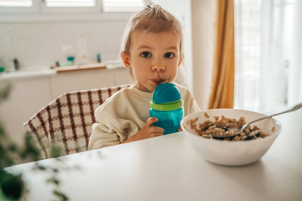 Peaceful little girl seated at the table with a bowl of cereal while sipping a healthy beverage - Fotoğraf, Görsel