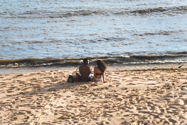 Salvador, Bahia, Brazil - December 12, 2021: Ένας άνδρας και μια γυναίκα κάθονται στην άμμο της παραλίας στην ύπαιθρο. Όμορφη ηλιόλουστη μέρα. Πόλη του Σαλβαδόρ, Βραζιλία. - Φωτογραφία, εικόνα