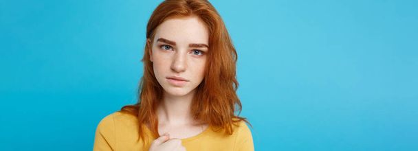 Headshot Portrait of tender redhead teenage girl with serious expression looking at camera. Caucasian woman model with ginger hair posing indoors.Pastel blue background. Copy Space - 写真・画像