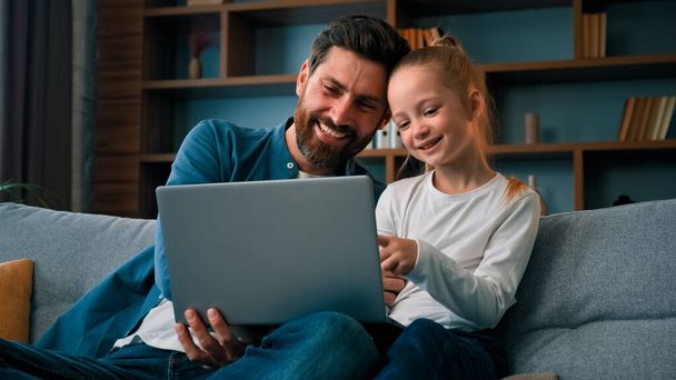 Cheerful father and little beautiful daughter girl sit on couch watch movies cartoons on internet spend time together at home having fun use laptop play games online modern tech usage parental control - Valokuva, kuva