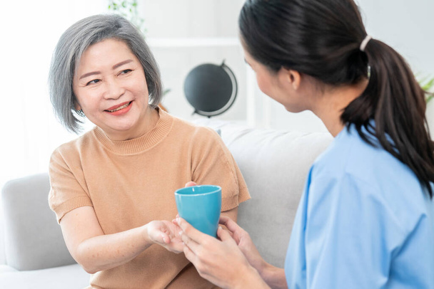 Female care taker serving her contented senior patient with a cup of coffee at home, smiling to each other. Medical care for pensioners, Home health care service. - Φωτογραφία, εικόνα