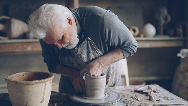 Doświadczony ceramista siwowłosy brodaty mężczyzna wygładza ceramiczny garnek mokrą gąbką. Kręcące się koło do rzucania, błotnisty stół roboczy i ręcznie robione clayware są widoczne. - Zdjęcie, obraz