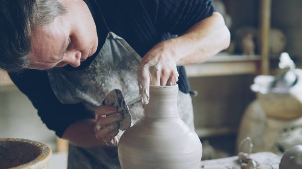 Close-up shot of skillful young potter molding ceramic vase from clay on throwing wheel while working in potters workshop. Shelves with handmade eathenware in background. - Foto, afbeelding