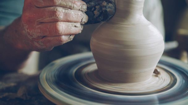 Elderly bearded craftsman is creating ceramic jar on potters wheel using professional tools. Workplace with handmade pots, beautiful vases and figures in background. - Fotó, kép
