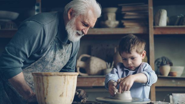 Concentrated young boy is molding clay into ceramic pot on spinning throwing wheel and his loving experienced grandfather is talking to him. Pottery and family tradition concept. - Фото, изображение