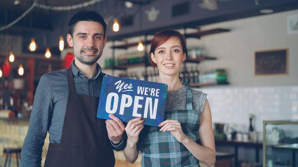 Pretty red-haired businesswoman cafe owner is holding yes we are open sign with her cheerful employee in apron standing near her. Successful start-up and people concept. - Photo, Image