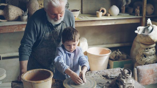 Young cute boy is making clay figure on throwing wheel while his caring grandfather experienced sculptor is standing near worktable and watching carefully. Family tradition concept. - Foto, Imagen