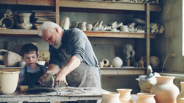 Professional potter is cutting ceramic pot from throwing wheel and his helpful little grandchild is bringing it to work table. Family members working together concept. - Photo, Image