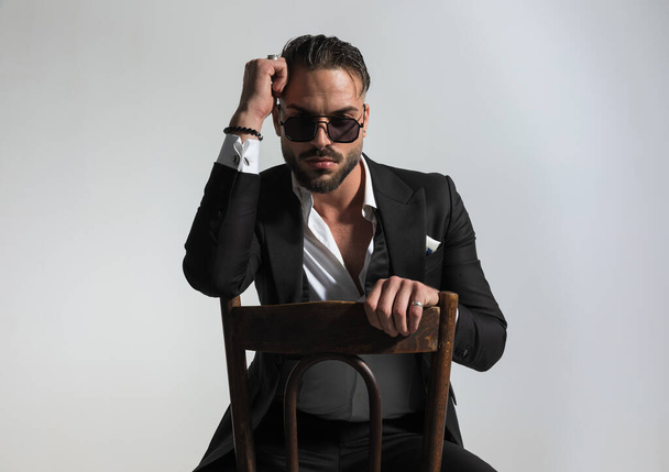 portrait of cool guy in black tuxedo holding elbow on old wooden chair and posing in a fashion manner on grey background  - Foto, imagen