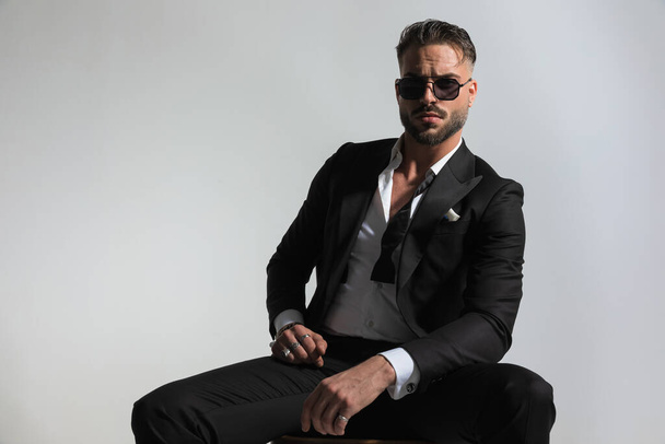 arrogant elegant groom wearing black tuxedo with open collar shirt and sunglasses holding arms in fashion pose and posing on grey background - Photo, Image