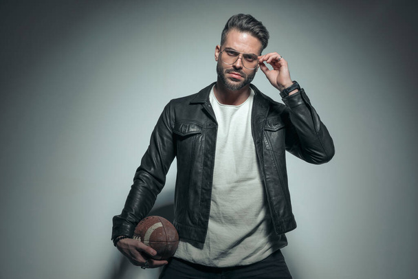 attractive casual man arranging his glasses while holding a rugby ball on his hip and posing with tough vibe - Fotó, kép