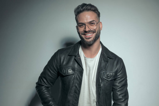 sexy young casual man giving a big and warm smile, wearing a black leather jacket and eyeglasses - Фото, изображение