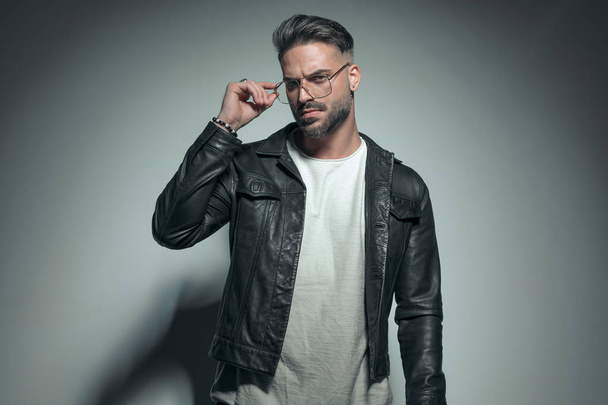 sexy fashion model arranging his glasses and looking at the camera with deep look on gray background - Photo, Image