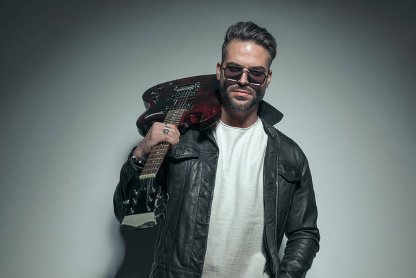 sexy rocker holding his electric guitar on his back and posing with cool sunglasses on gray background  - Photo, Image