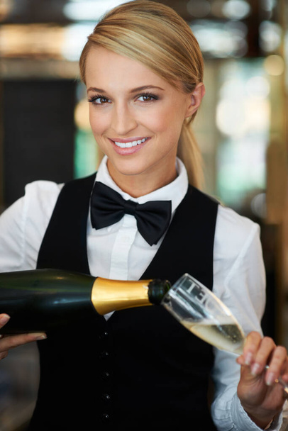 Давайте выпьем за вас. Привлекательная официантка наливает шампанское в бокал шампанского, улыбаясь в камеру. - Фото, изображение